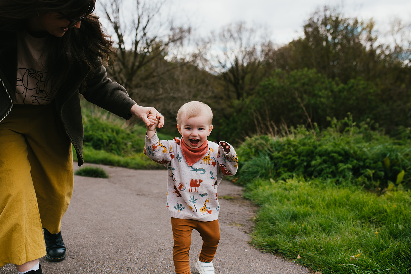 A young child walking in a park. They are holding an adult's had and smiling towards the camera.
