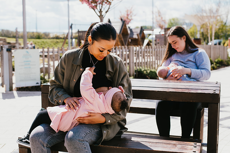 Two woman sat outdoors at a picnic bench. Both are breastfeeding babies