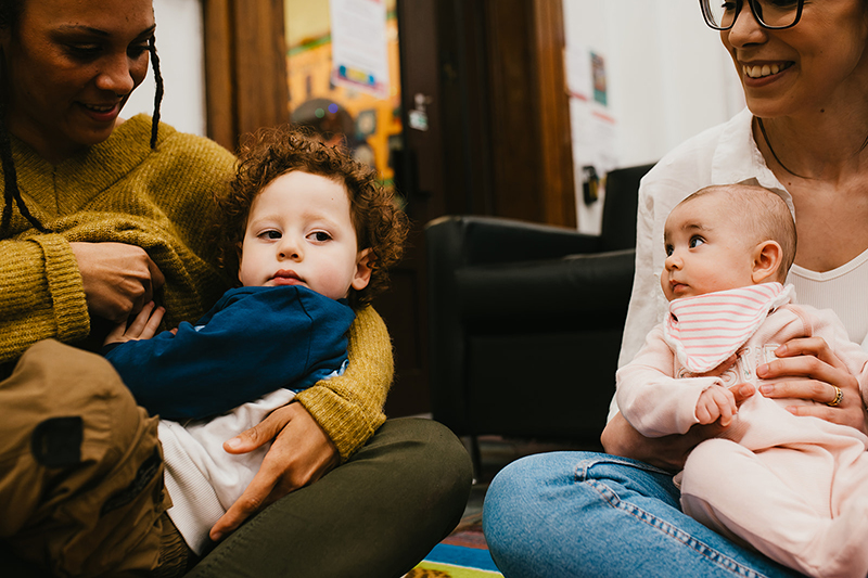 A women holding a toddler sat next to a woman holding a baby
