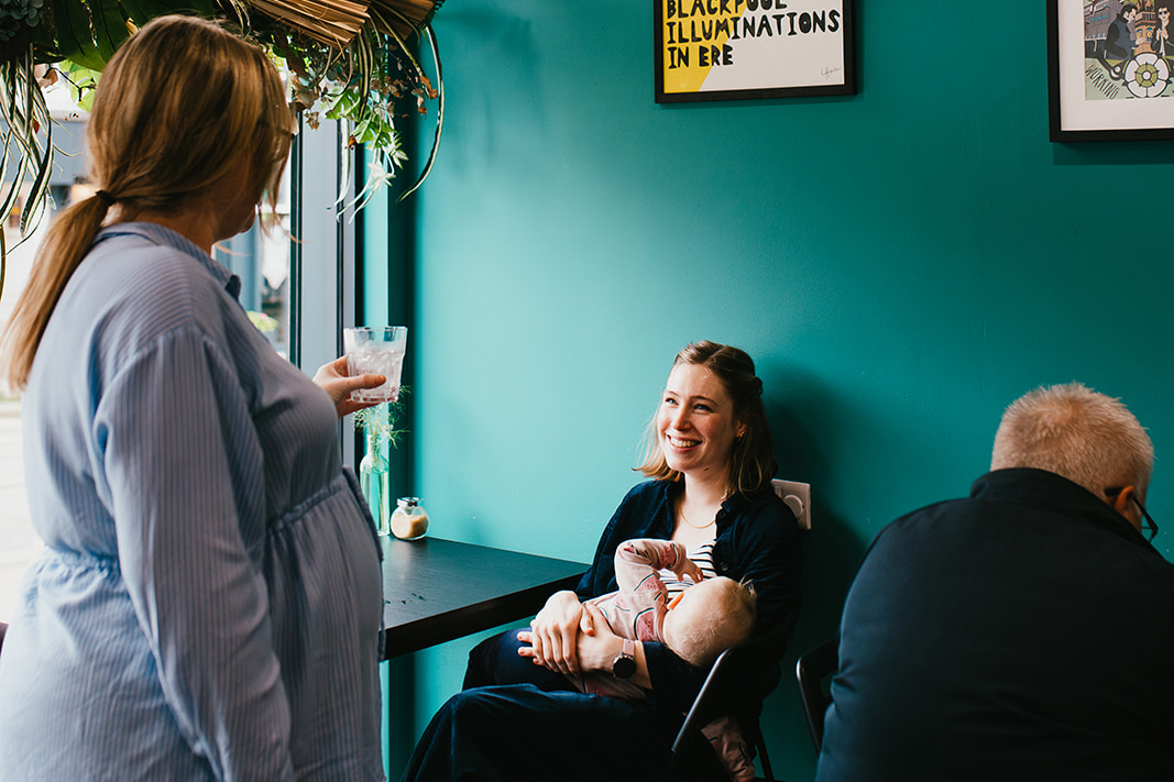 A woman sat breastfeeding. Someone is offering her a drink